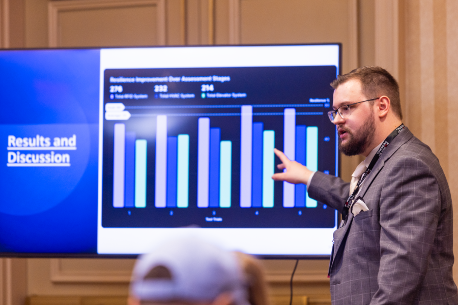 A man stands in front of a screen and presents to a large group about research depicted on the screen in the form of a bar graph.