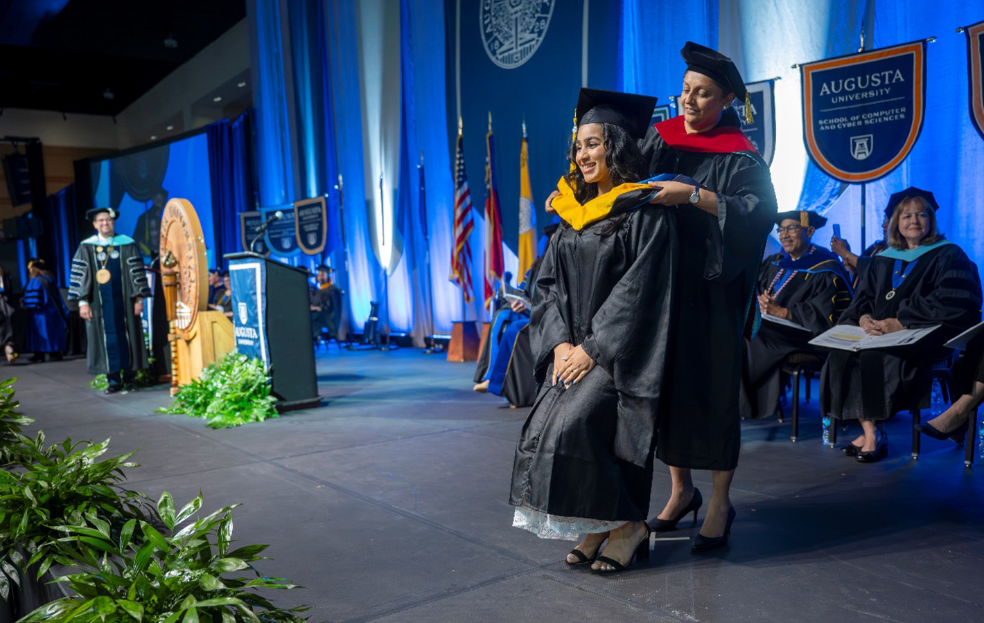 Person wearing cap and gown places a hood on a female student in cap and gown.